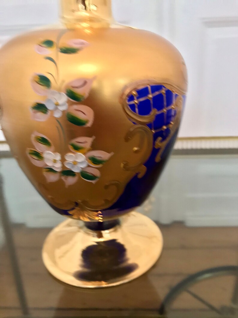 May include: A cobalt blue glass vase with gold accents and floral designs. The vase has a wide base and a narrow neck.