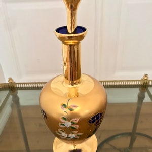 May include: A gold glass decanter with a blue glass stopper and floral painted details. The decanter has a round body and a wide base.