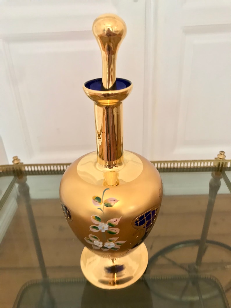 May include: A gold glass decanter with a blue glass stopper and floral painted details. The decanter has a round body and a wide base.