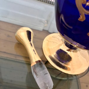 May include: A gold bottle stopper with a clear glass insert. The stopper is on a glass surface.