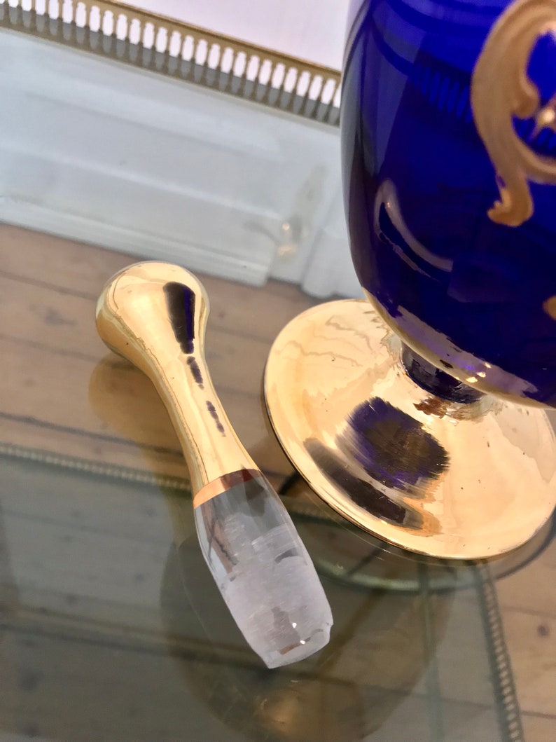May include: A gold bottle stopper with a clear glass insert. The stopper is on a glass surface.