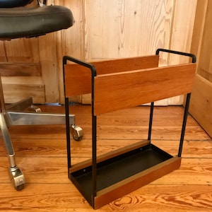 Puede incluir: Un soporte para paraguas de metal negro con parte superior e inferior de madera. El soporte tiene dos estantes para guardar paraguas.