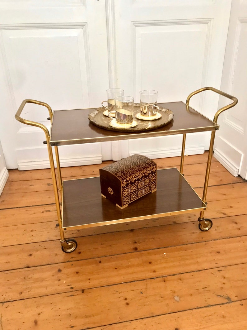 Puede incluir: Un carro de servicio de metal dorado con dos niveles. El nivel superior tiene una superficie de madera marr&oacute;n y una bandeja de metal dorado con tres tazas de vidrio. El nivel inferior tiene una superficie de madera marr&oacute;n y una peque&ntilde;a caja de madera con un patr&oacute;n decorativo. El carro tiene cuatro ruedas.