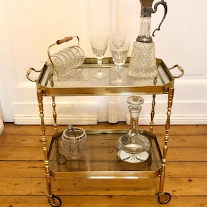 May include: A two-tiered gold-toned drinks trolley with glass shelves. The top shelf holds two wine glasses, a crystal jug, and a glass coaster set. The bottom shelf has a glass decanter and a small glass bucket. The trolley has wheels.