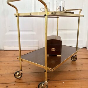Puede incluir: Un carro de servicio de metal dorado con dos estantes de madera. El carro tiene cuatro ruedas y un asa. Hay una peque&ntilde;a caja de madera en el estante inferior.