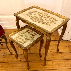 Puede incluir: Dos mesas nido antiguas. Las mesas tienen un borde dorado y tapas color crema con un diseño floral dorado. Las patas curvas son de madera. Las mesas están sobre un suelo de madera.