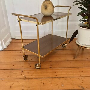 Puede incluir: Un carrito de servicio de metal dorado con dos niveles y cuatro ruedas. El carrito tiene una tapa de madera marr&oacute;n y un jarr&oacute;n de cer&aacute;mica marr&oacute;n en el nivel superior.