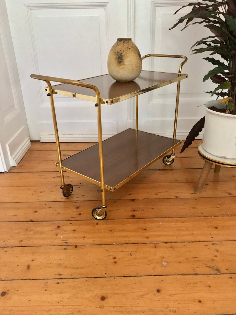 Puede incluir: Un carrito de servicio de metal dorado con dos niveles y cuatro ruedas. El carrito tiene una tapa de madera marr&oacute;n y un jarr&oacute;n de cer&aacute;mica marr&oacute;n en el nivel superior.