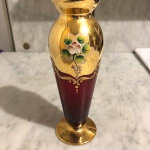 May include: A small, red glass vase with gold accents and a painted floral design. The vase has a round base and a narrow neck.