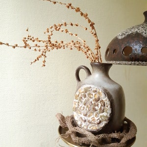 Puede incluir: Un jarrón de cerámica con un diseño texturizado blanco y marrón, lleno de tallos de flores marrones secas. El jarrón está sentado en un soporte dorado con una base de arpillera.