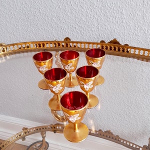 以下が含まれることがあります： 金色の縁取りが施された赤いガラス製のショットグラス6個セット。白い花柄のデザインが施されています。グラスは、金色のトレイの上に三角形に配置されています。