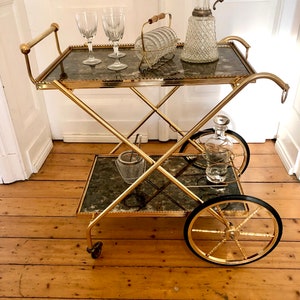 Puede incluir: Un carrito de bar dorado con dos estantes de vidrio y una tapa negra. El carrito tiene cuatro ruedas y un diseño decorativo en los lados. Hay tres copas de vino, una botella de licor y una botella de vino en el carrito.