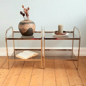 Puede incluir: Dos mesas auxiliares de metal dorado con estantes de vidrio. La mesa de la izquierda tiene un jarrón de cerámica marrón con flores secas en el estante superior y un libro en el estante inferior. La mesa de la derecha tiene un plato blanco con una vela encima.