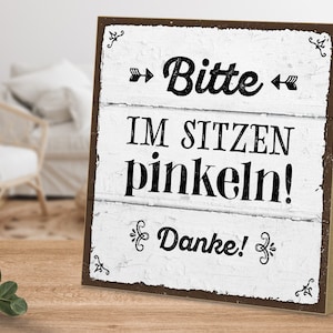 Könnte beinhalten: Ein weißes Holzschild mit schwarzem Text, der "Bitte im sitzen pinkeln! Danke!" lautet. Das Schild hat einen braunen Holzrahmen.
