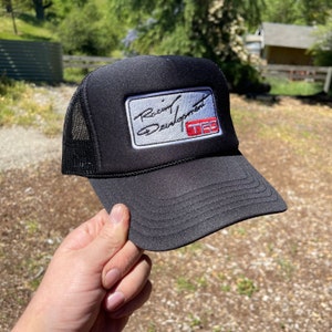 May include: A black trucker cap featuring a rectangular patch on the front panel. The patch displays the text Racing Development in black script and the letters TRD in red within a white rectangular border. The cap has a mesh back and a curved brim.