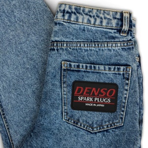 May include: Acid-wash blue denim jeans featuring a rectangular black patch on the back pocket. The patch displays the red text DENSO, white text SPARK PLUGS, and white text MADE IN JAPAN, all enclosed within a thin red rectangular border.