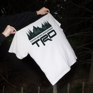 Peut inclure: Un t-shirt blanc avec une impression graphique noire représentant une silhouette de chaîne de montagnes au-dessus du texte TOYOTA RACING DEVELOPMENT et des grandes lettres TRD en noir. Le motif est placé sur le haut du tissu blanc.