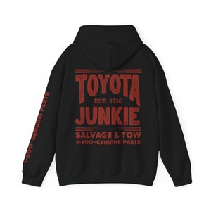 May include: A black hooded sweatshirt featuring red distressed-style text on the back that reads TOYOTA JUNKIE EST. 1936 SALVAGE & TOW 1-800-GENUINE PARTS. The left sleeve also displays the red text 1-800-GENUINE PARTS running vertically down the arm.