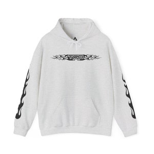 May include: A light grey heathered hoodie featuring a black graphic of the text TRD surrounded by flame patterns on the chest. The long sleeves also display black flame designs. The garment includes a hood with white drawstrings and a front pocket.