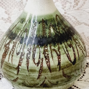 May include: A ceramic vase with a narrow neck and a bulbous body. The neck is a matte off-white, while the body features a glossy green glaze with dark brown, vertical, textured brushstroke patterns and a horizontal dark band around the centre.