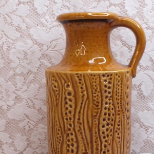 May include: A tall, brown ceramic vase with a handle, featuring a textured, bark-like design. The vase is accompanied by a bundle of dried wheat. The vase is approximately 25 cm tall.