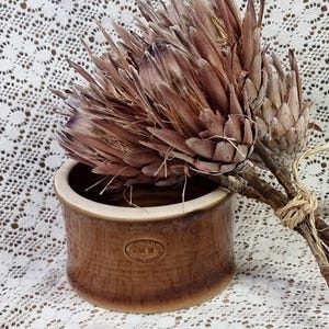 May include: A brown ceramic pot with a textured surface and a circular logo that says "M.H. Pottery" sits on a white lace tablecloth. A bouquet of dried protea flowers with brown stems is in the pot.