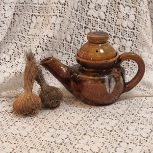 Puede incluir: Una tetera de cerámica marrón con tapa y un esmalte marrón y blanco. La tetera tiene un pico y una asa. La tetera está sobre un mantel de encaje blanco.