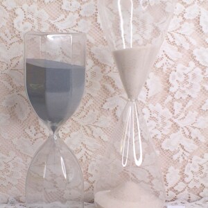 May include: Two clear glass hourglasses, one with grey sand and the other with white sand. The hourglasses are on a white lace surface. The hourglass on the left has a hexagonal base. The hourglass on the right has a rounded shape.