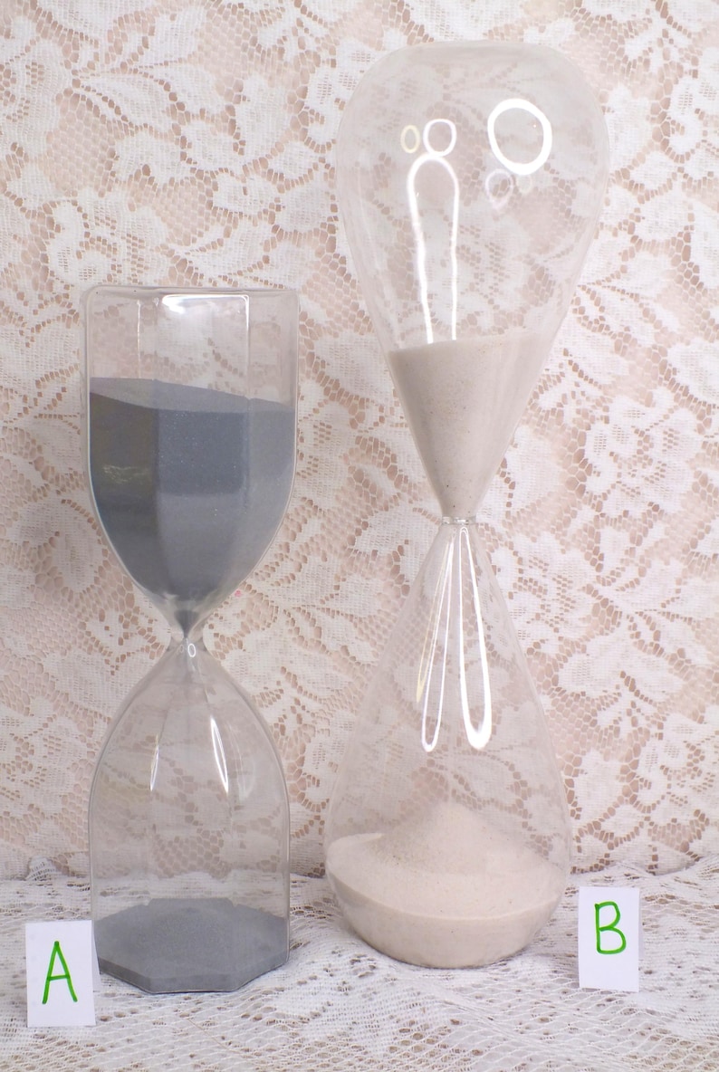 May include: Two clear glass hourglasses, one with grey sand and the other with white sand. The hourglasses are on a white lace surface. The hourglass on the left has a hexagonal base. The hourglass on the right has a rounded shape.