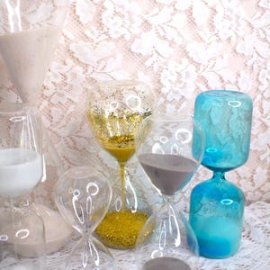 May include: A selection of glass hourglasses in different sizes and colours. Some contain gold glitter, grey sand, and white sand. The hourglasses are displayed on a white lace surface.