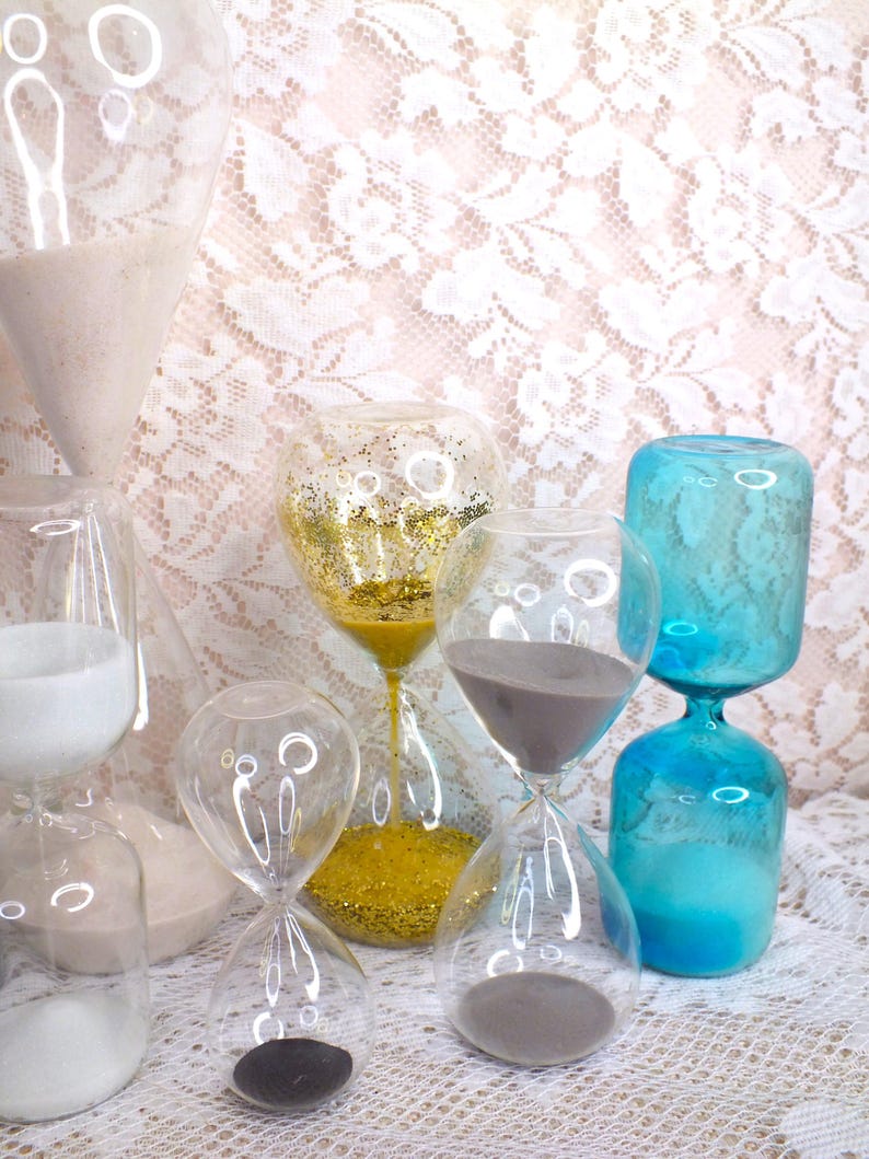 May include: A selection of glass hourglasses in different sizes and colours. Some contain gold glitter, grey sand, and white sand. The hourglasses are displayed on a white lace surface.