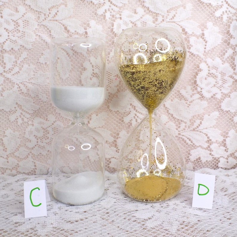May include: Two glass hourglasses, one with white sand and the other with gold glitter. The hourglasses are clear with a bulbous shape, sitting on a white lace surface. The white sand is in the process of flowing from the top to the bottom.