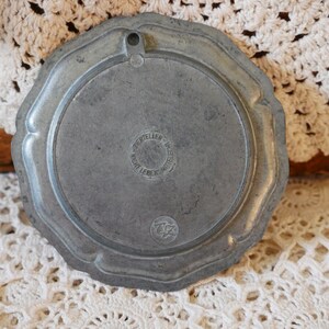 May include: A round, silver-colored metal plate with a raised edge and a circular design in the center. The plate has a hole at the top for hanging. The back of the plate has the text "ZERTIELLER" and "NICHT LEBENSMITTEL" in a circle.