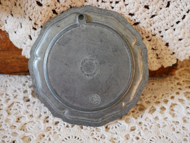May include: A round, silver-colored metal plate with a raised edge and a circular design in the center. The plate has a hole at the top for hanging. The back of the plate has the text "ZERTIELLER" and "NICHT LEBENSMITTEL" in a circle.