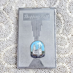 May include: A rectangular silver-toned metal shopping list case with a textured cross-hatch pattern. The top features the engraved text Shopping List above a central enamel emblem depicting St. Paul's Cathedral in Melbourne with blue sky.