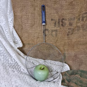 Op de afbeelding: Een metalen zeef met een blauwe handgreep en een groene appel erin. De zeef ligt op een wit kantkleed en een bruine jute achtergrond.