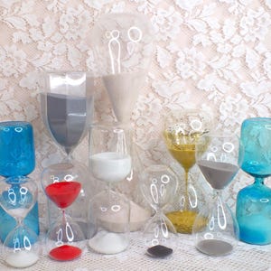 Puede incluir: Una colección de relojes de arena de vidrio transparente con arena de colores. Los relojes de arena presentan diferentes formas y colores, incluyendo azul, rojo, dorado y gris. Algunos tienen bases decorativas, y la arena en el interior varía en color.