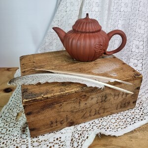 Pode incluir: Uma chaleira de terracota com um design floral está sobre uma caixa de madeira com uma pena em cima.