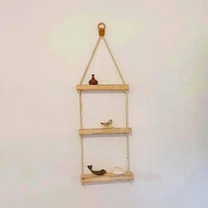 May include: A three-tiered hanging shelf crafted from light wood and rope. The shelf displays decorative items: a brown vase, a small figurine, a wooden whale, and a seashell. The shelf is suspended against a white wall, offering a rustic aesthetic.