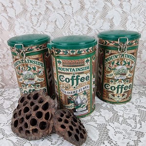 May include: Three cylindrical metal coffee canisters with green lids and latch closures, featuring vintage-style labels that read Mountainside Coffee, Mocha Java, and Full-Bodied Flavor, accompanied by two dried, dark brown lotus seed pods.