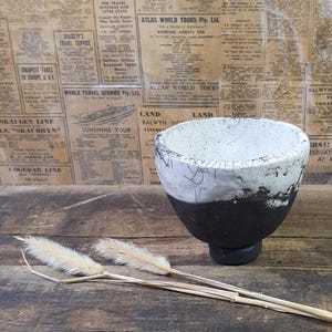 May include: A handcrafted ceramic bowl with a white and black glaze. The bowl has a rounded shape and a footed base. The background features vintage newspaper clippings.
