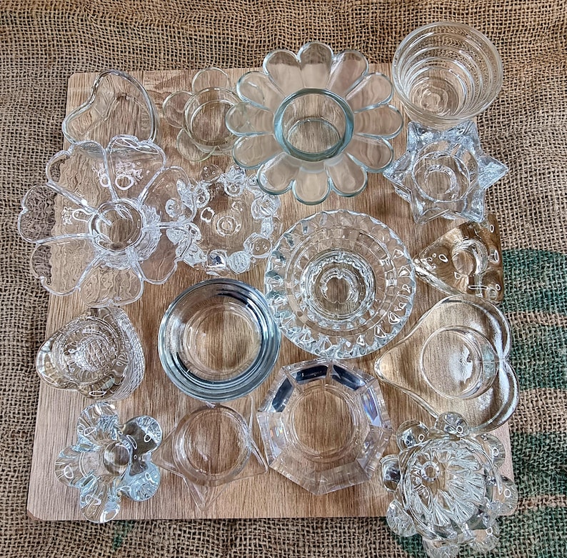 May include: A collection of clear glass candle holders and small dishes in various shapes, including flowers, stars, and geometric designs. The items are arranged on a light brown wooden board.