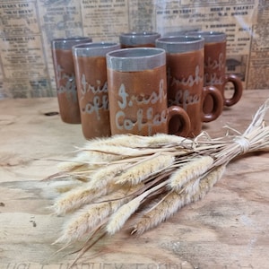 Puede incluir: Conjunto de seis tazas de cerámica marrón con borde gris y las palabras "Irish Coffee" escritas en blanco en la parte delantera. Las tazas están dispuestas en fila sobre una superficie de madera con un manojo de hierba seca delante de ellas.