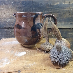 May include: A rustic brown ceramic jug with a handle and spout, adorned with dark brown and black glaze patterns. Three decorative dried plant ornaments with a spiky texture sit beside it, all on a wooden surface.