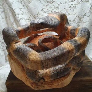 May include: A wooden sculpture of a snake coiled around itself. The snake is painted with brown, black, and tan colours. The sculpture is set on a wooden base, and the snake's head is raised.