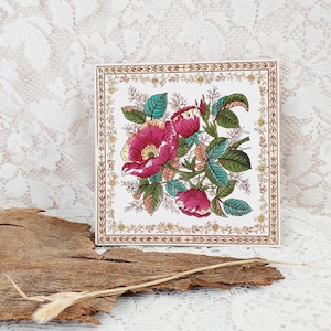 Könnte beinhalten: Eine weiße Keramikfliese mit einem floralen Design. Die Fliese zeigt einen Strauß rosa Rosen mit grünen Blättern und Stielen. Die Blumen sind von einem goldenen und braunen Rand umgeben.