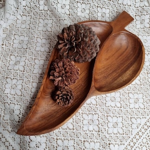 Puede incluir: Un plato de servir de madera con forma de hoja con dos compartimentos separados. El plato es de madera marrón oscura y tiene un acabado natural. Tres pequeñas piñas se colocan en el plato.