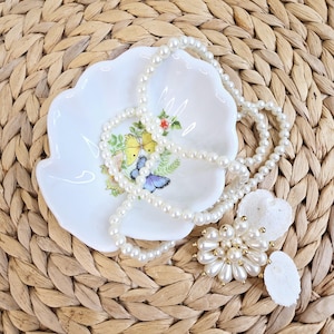 May include: A white ceramic trinket dish with a floral design featuring butterflies. A pearl necklace and a pearl brooch are displayed on the dish.