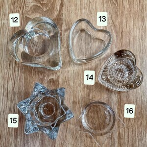 May include: A collection of clear glass candle holders in various shapes, including hearts and stars. The candle holders are displayed on a wooden surface. The shapes include a double heart, a single heart, and a star.