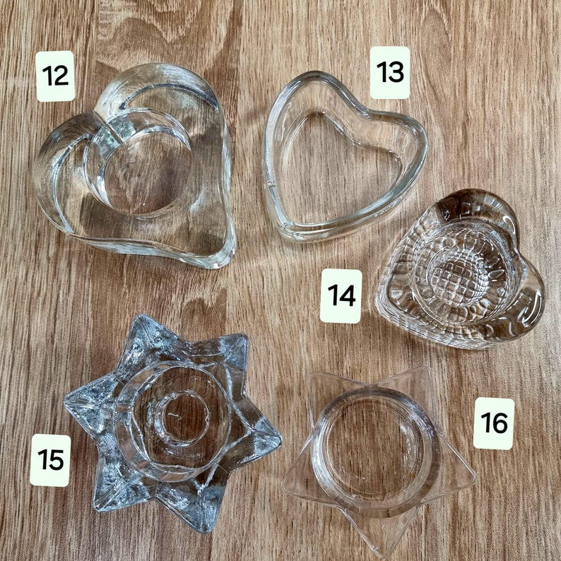 May include: A collection of clear glass candle holders in various shapes, including hearts and stars. The candle holders are displayed on a wooden surface. The shapes include a double heart, a single heart, and a star.